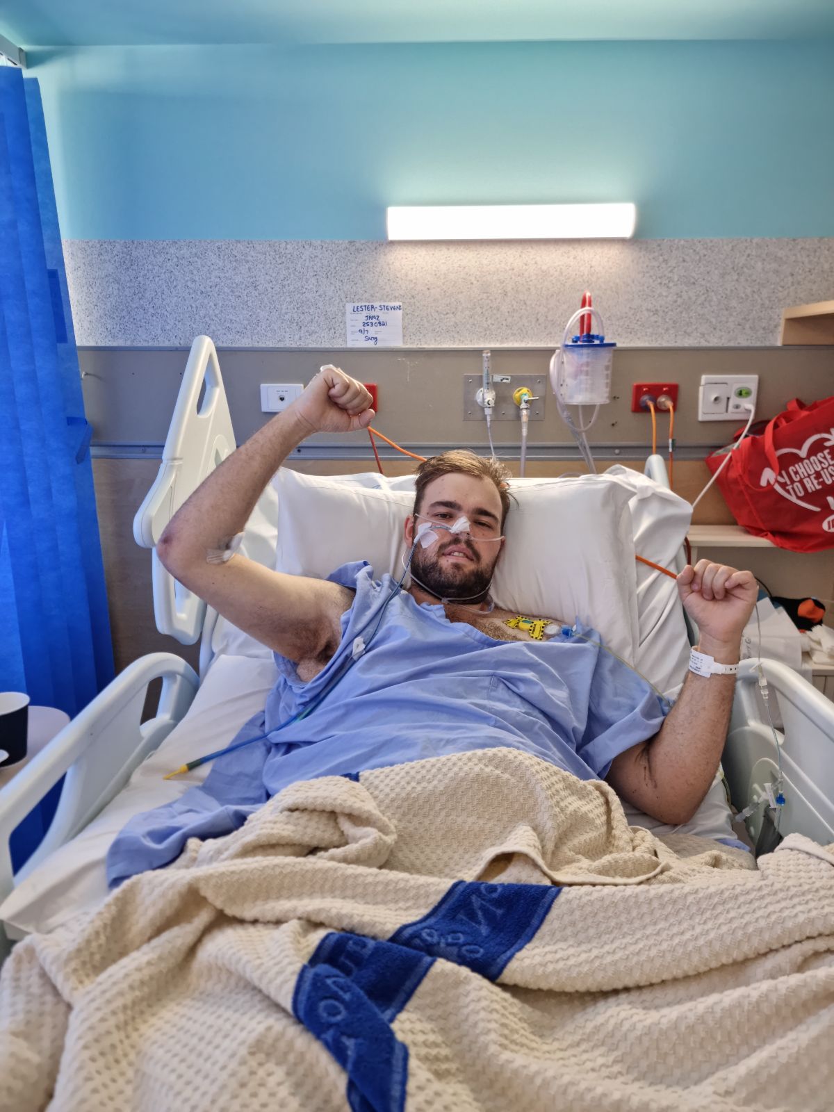 Jaymz Lester-Stevens is lying in a hospital bed, with a blue hospital gown with hands up. Medical equipment all around.