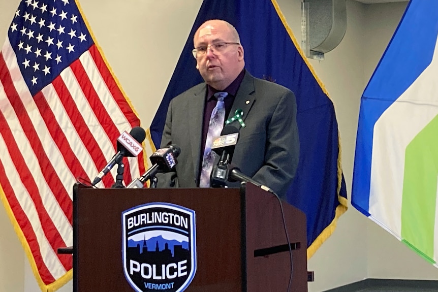 Tom Curran in a suit speaks in front of flags at a lectern at a Burlington Police press conference