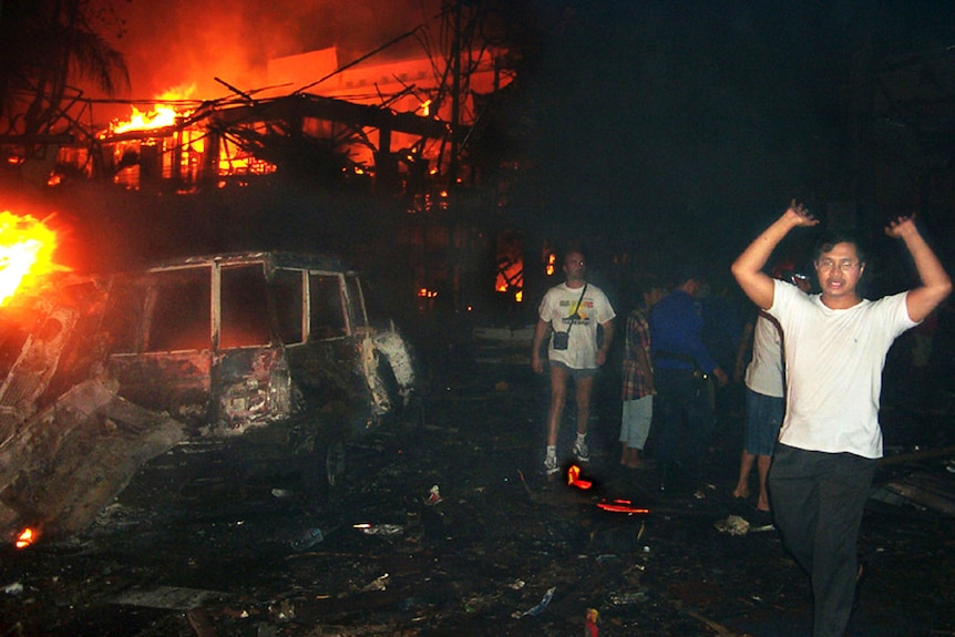 A group of people with their hands in the air leave the wreckage of a building on fire.