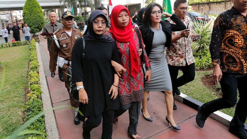 A woman in a red headcarf walks flanked by two other women and reporters and police on her way to court.