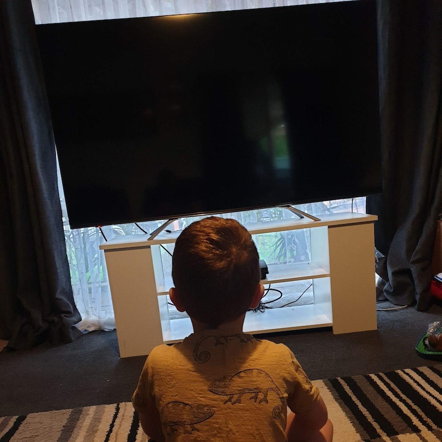 back head of a child sitting cross-legged on the ground in a small room watching TV