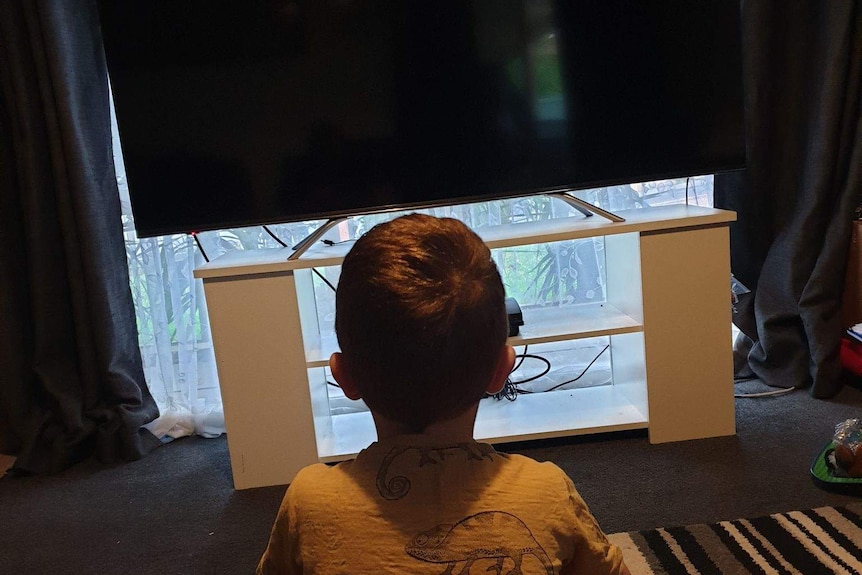 A child sitting cross-legged on the ground in a small room watching TV