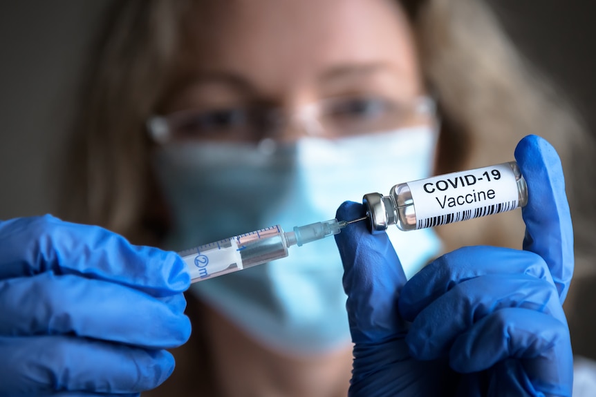 A woman wearing a surgical mask and blue gloves holds up a COVID vaccine vial and a syringe.