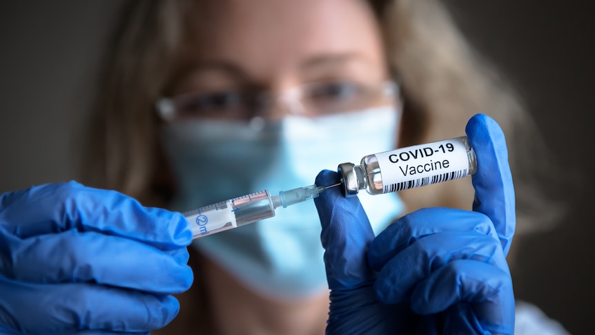 A woman wearing a surgical mask and blue gloves holds up a COVID vaccine vial and a syringe.