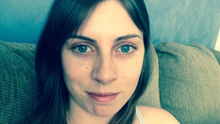 A young woman sits on a couch staring at camera, half smiling, with blue eyes and brown hair.