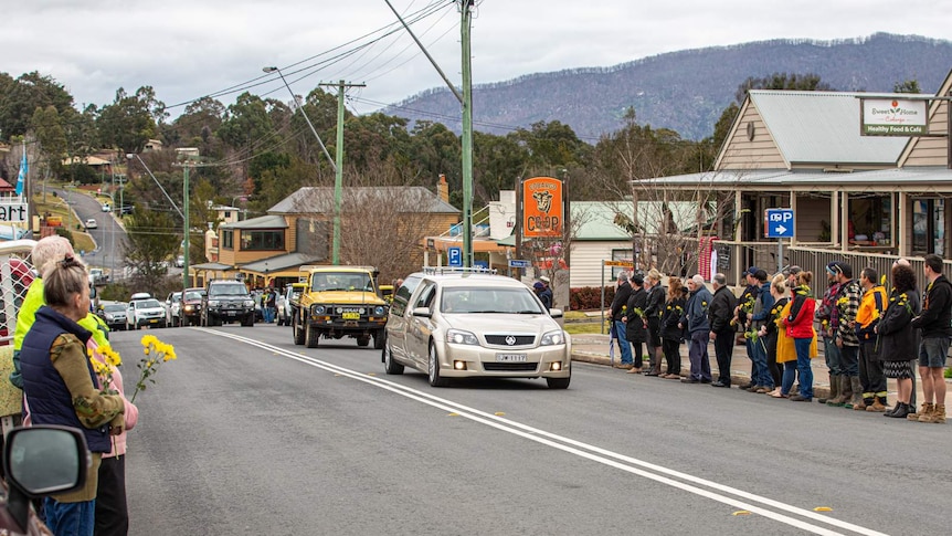 Men and women stand along street of small country town holding yellow flowers, as a hearse and yellow ute drive past.