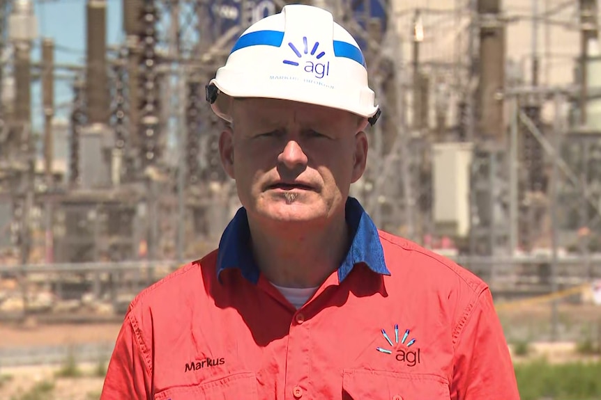 A man wearing an orange top and white hard hat stands in front of a power station with a serious expression