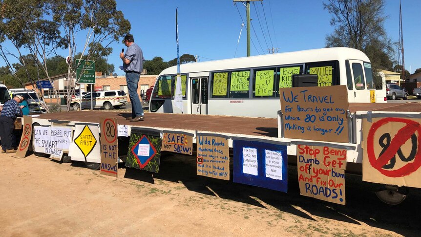 Man stands on a stage decorated with painted signs protesting the speed limit reduction along Robinvale-Sea Lake Road.