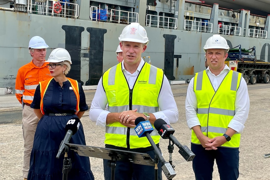 A politician in high-vis and a hard hat stands with his hands clasped, speaking to the media on a dock.