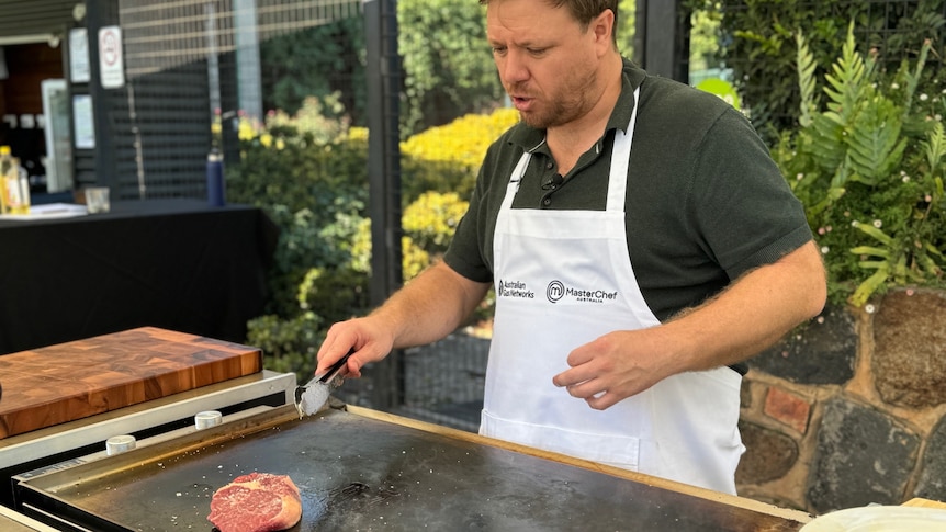 A man wearing a black polo shirt and white apron stands behind a barbecue cooking a steak.