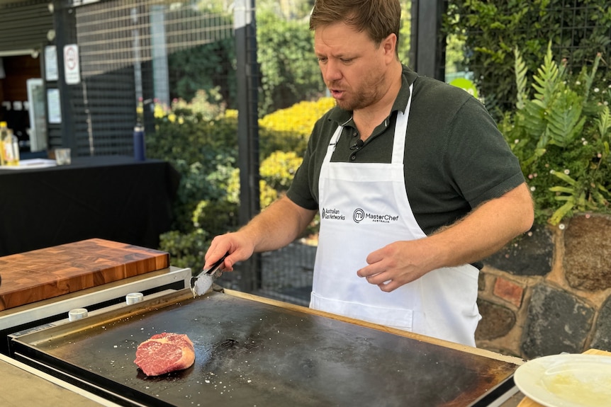 A man wearing a black polo shirt and white apron stands behind a barbecue cooking a steak.