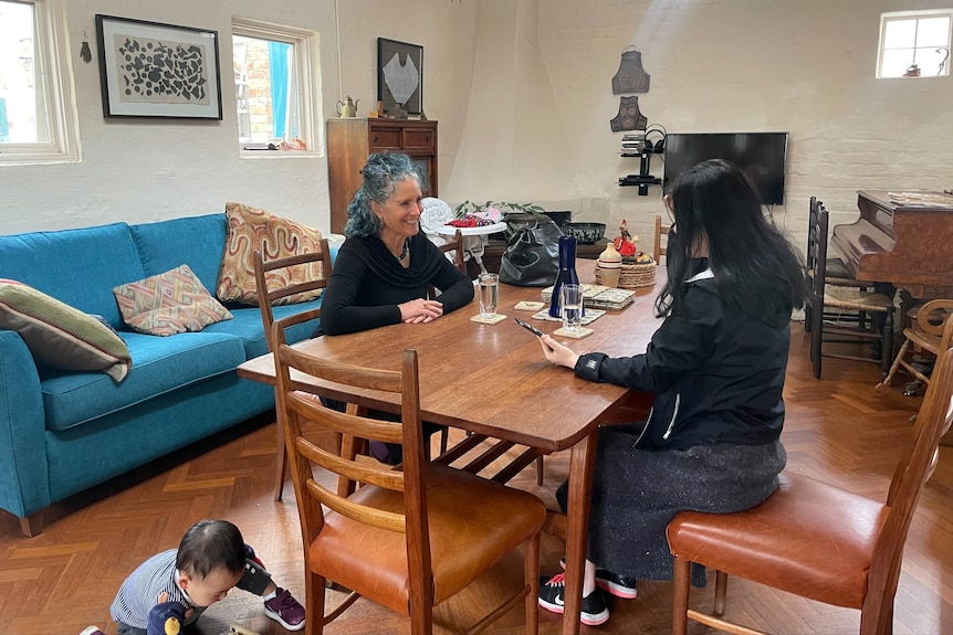 A woman with graying, curly hair sits across from a young woman in a living room while a child plays on the floor. 