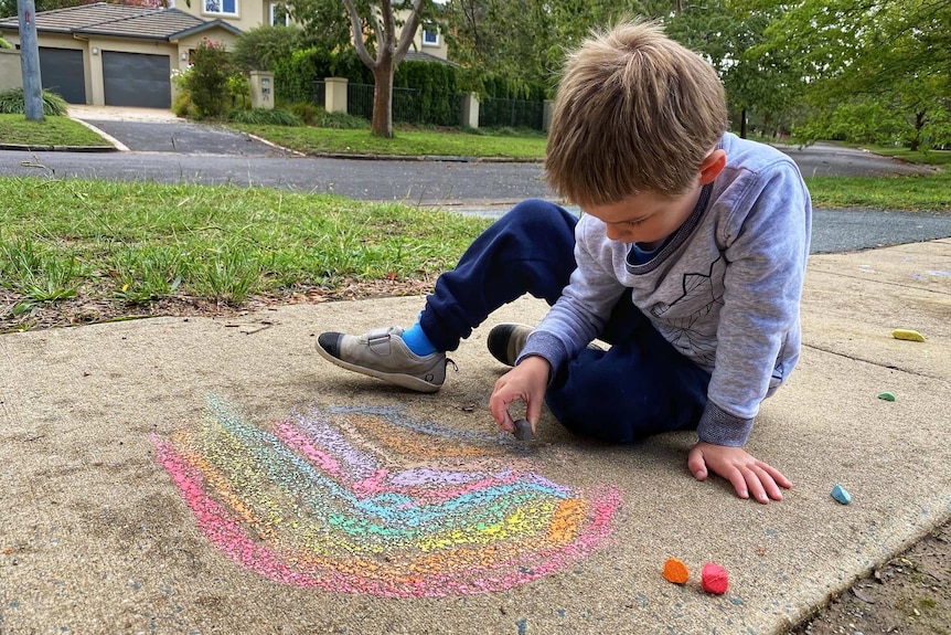 A boy draws a rainbow in chalk on the footpath