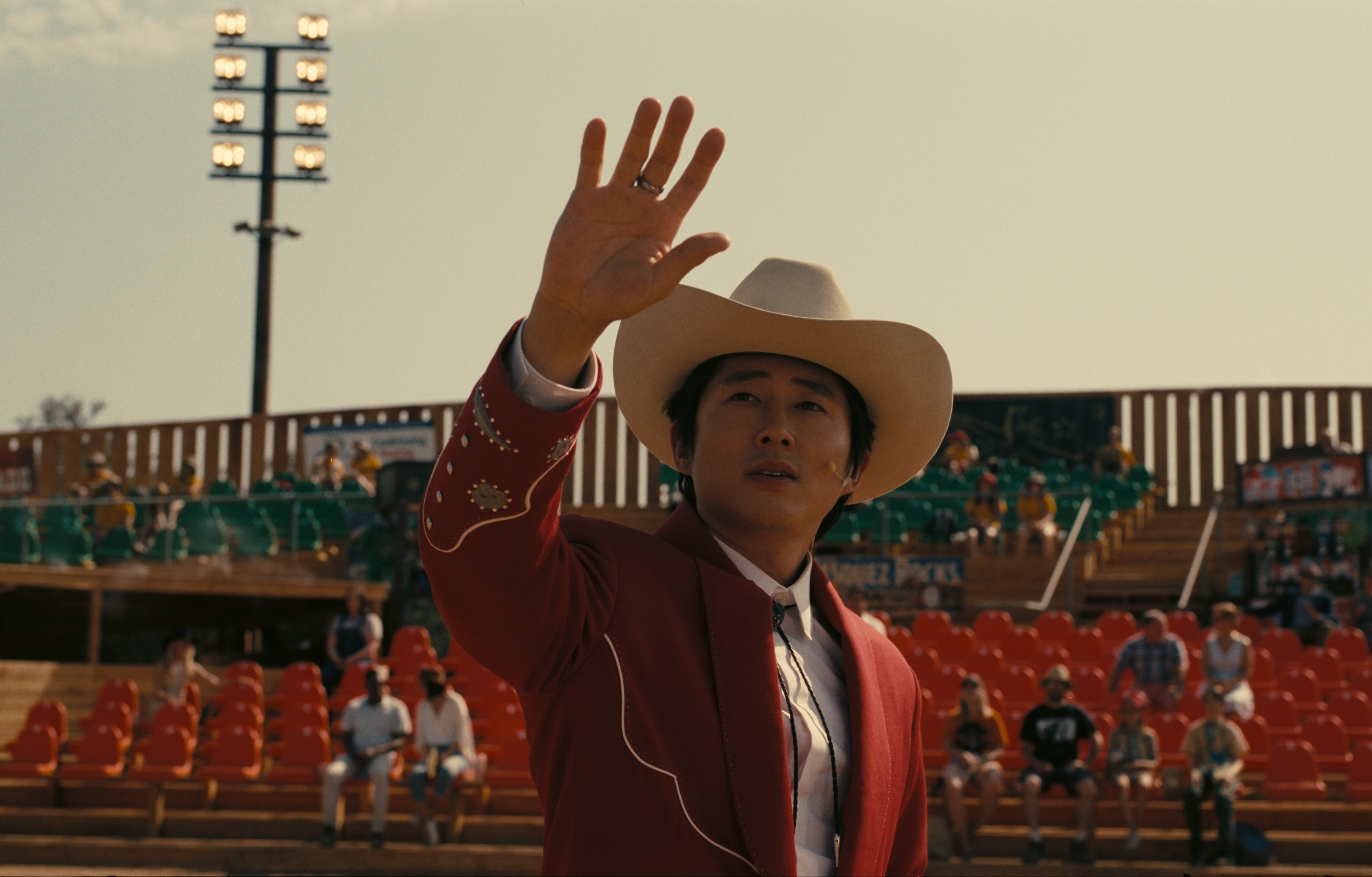 A man wearing a cowboy hat and a red jacket stands with one hand raised in an outdoor stadium
