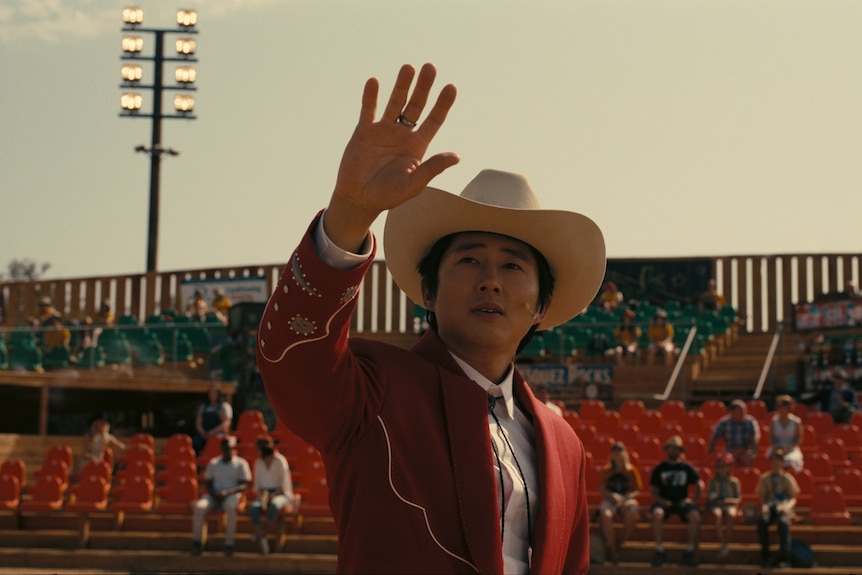 A man wearing a cowboy hat and a red jacket stands with one hand raised in an outdoor stadium