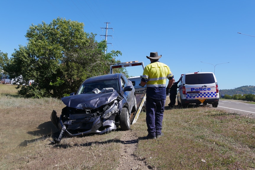 A man in high-vis stands on the side of a road in front of a trailer with a smashed-up car on it.