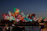 The Sydney Opera House is lit up with Indigenous artwork. It is twilight and onlookers can be seen taking in the view