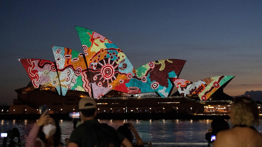 The Sydney Opera House is lit up with Indigenous artwork. It is twilight and onlookers can be seen taking in the view