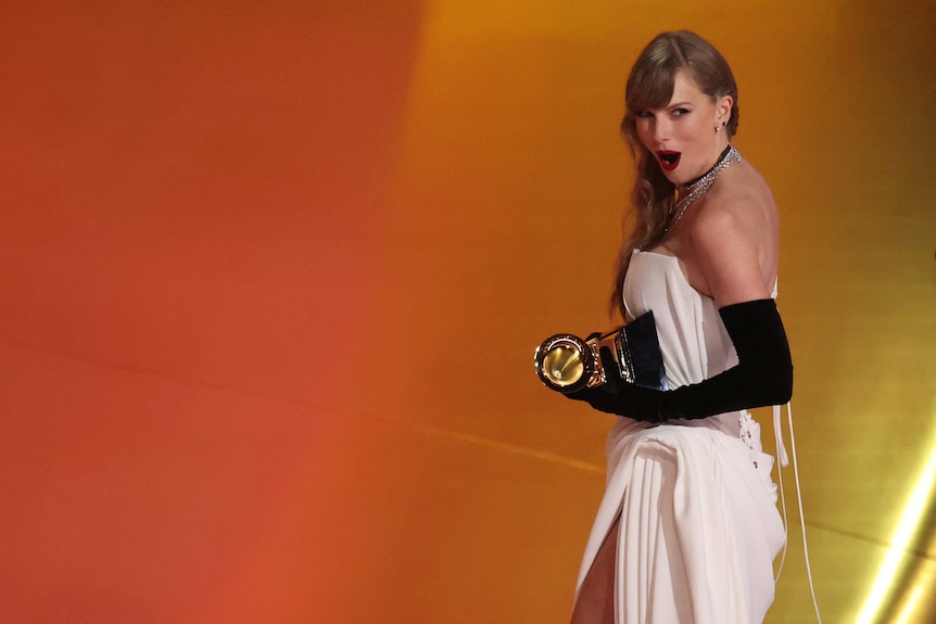 Taylor Swift with a shocked expression holding a Grammy and wearing a white dress.