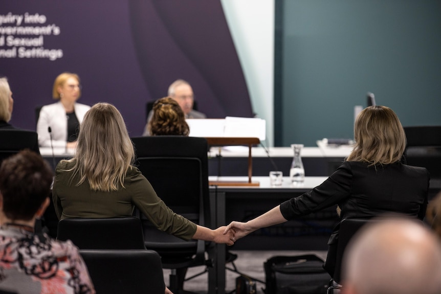 Two seated women hold hands in a public meeting, seen from behind.