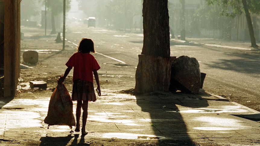 back of barefoot young girl carrying trash bag walking down rubbish strewn road.