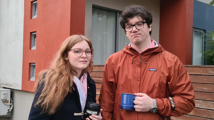 Sara and Alan stand in the street dressed in warm jackets with mugs in their hands.