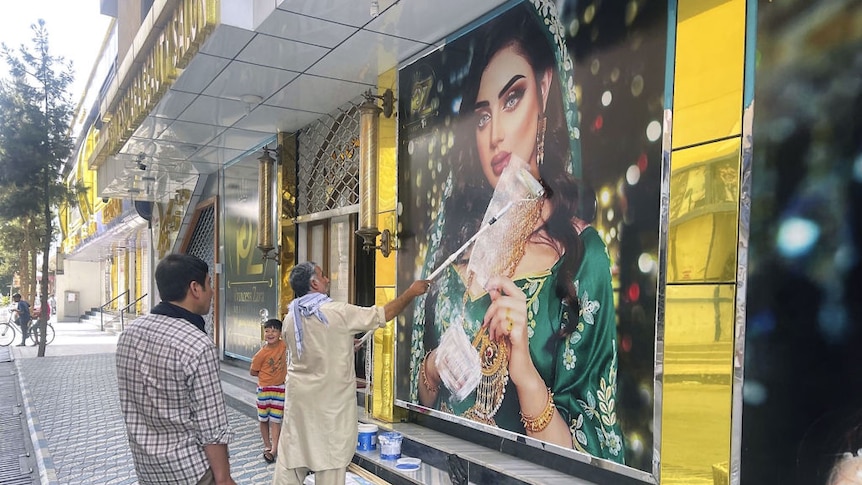 A worker outside a beauty salon paints over a large poster of a woman on the wall in Kabul watched over by two young boys.