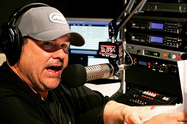 A man wearing a cap and t shirt talks into a microphone while seated in a radio broadcast studio