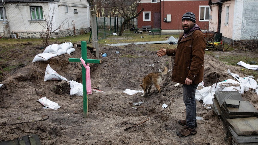 A white man in winter clothes stands next to a large crucifix near a recently dug backyard grave, amid sandbags and a dog.