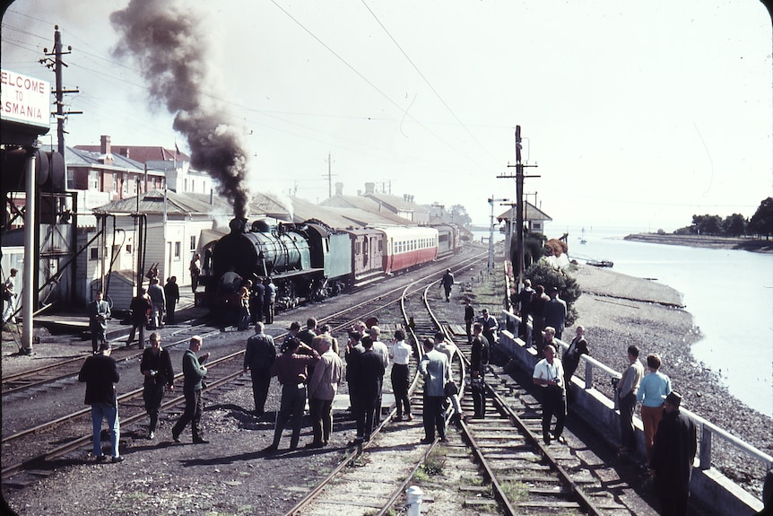 People gather by the rails as a steam train prepares to leave a station by a river