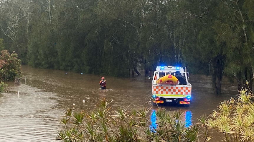 A person in high-vis wades through floodwater, near where an emergency vehicle is parked.