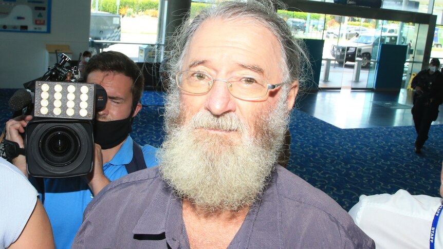 A bearded older man wearing glasses is followed by media