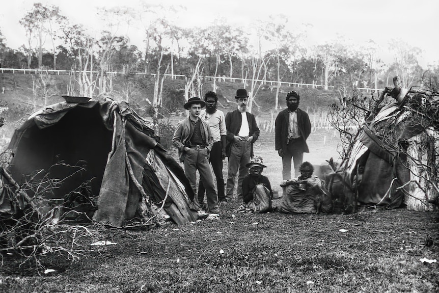 Indigenous and non-Indigenous people stand around huts.