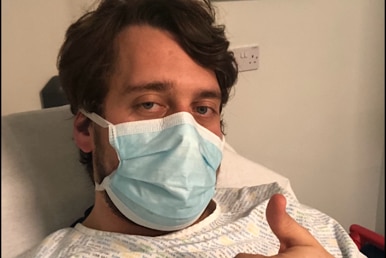 A man sits in a hospital gown and face mask, with a thumbs up to the camera. 