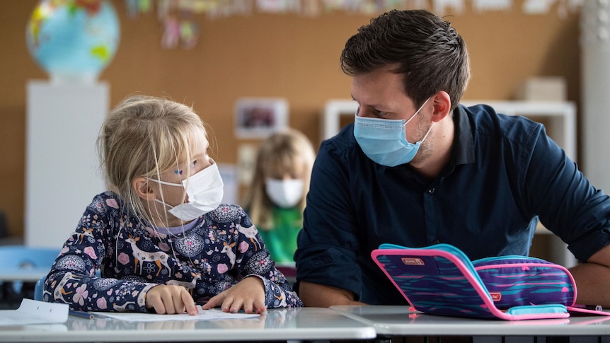 A male teacher and female student sit together in school classroom wearing face masks 