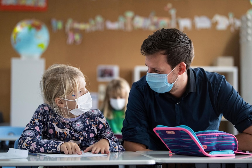 A male teacher and female student sit together in school classroom wearing face masks 