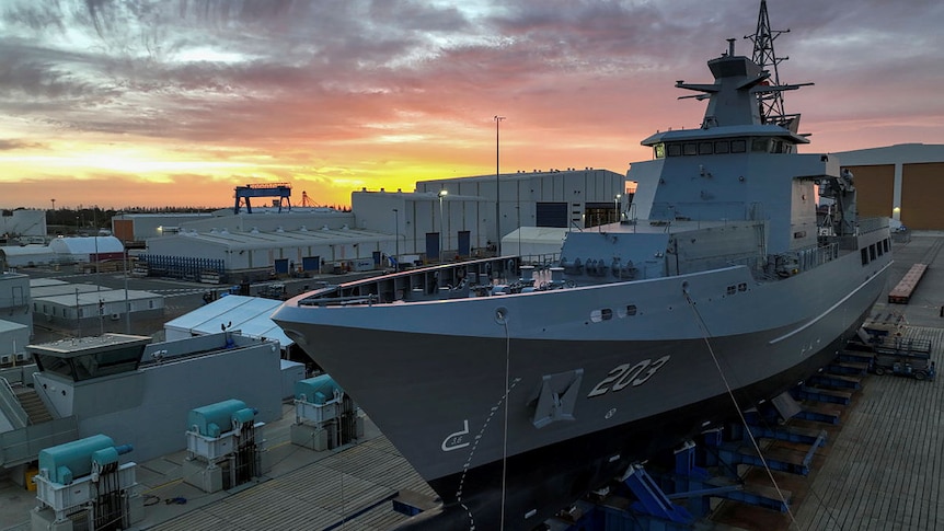 A large naval ship sits on a dock at sunset. 