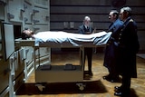 A young man's body lies in a morgue, with three men in military uniform looking over him