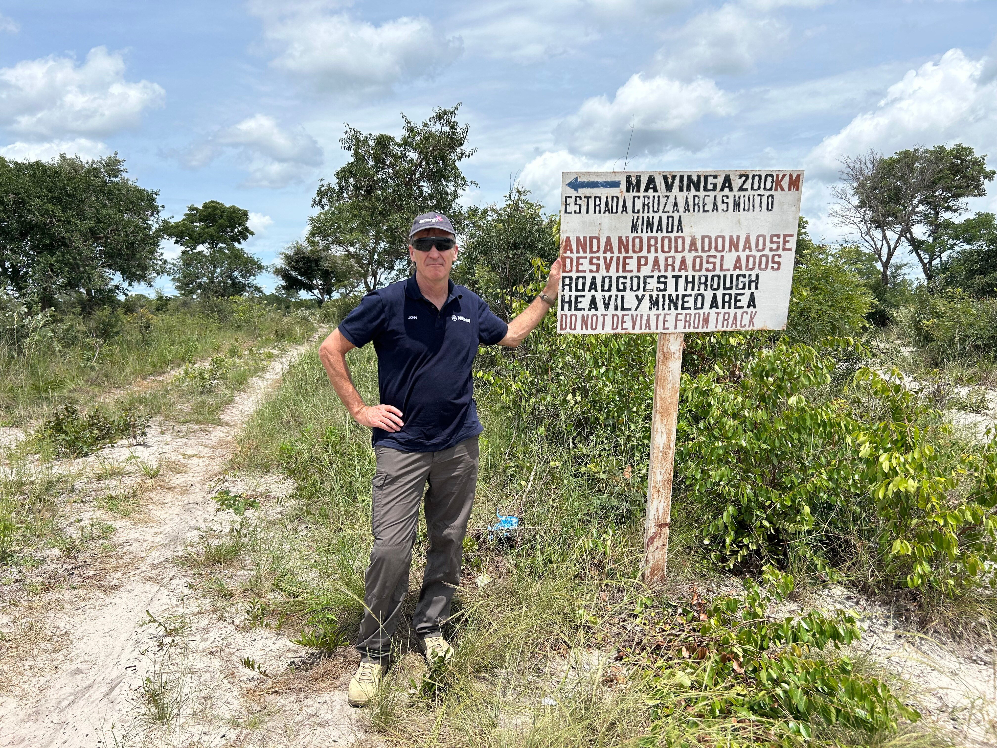 A man stands a trail in front ofa sign warning about landmines in different languages.  