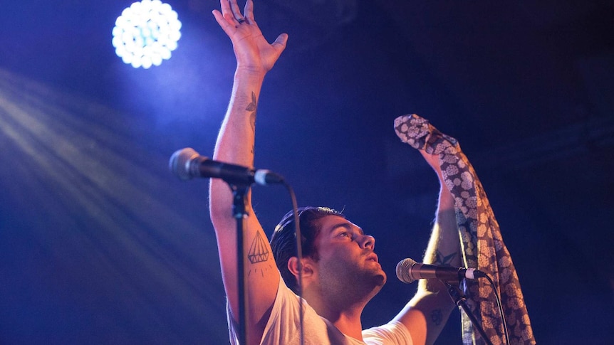 Dan Sultan  performing on stage with hands in the air and a light shoning above his head. Blue background, looking ahead