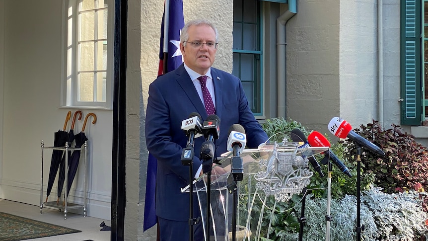 Scott Morrison standing at a podium in front of two australian flags in front of a sandstone house