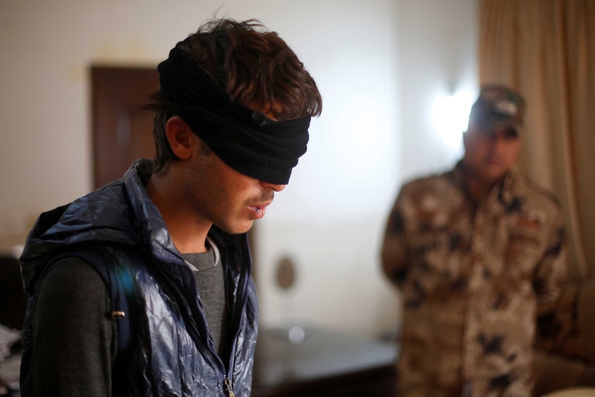 A boy stands blindfolded with a bleeding lip as an Iraqi soldier watches him.