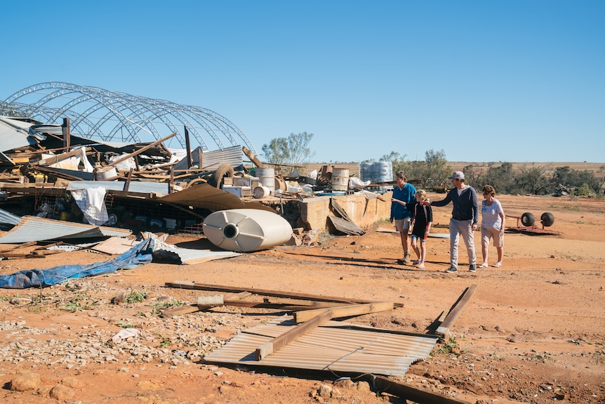A man, woman and their two children stand on red dirt on a farm next to a shed destroyed by a cyclone.