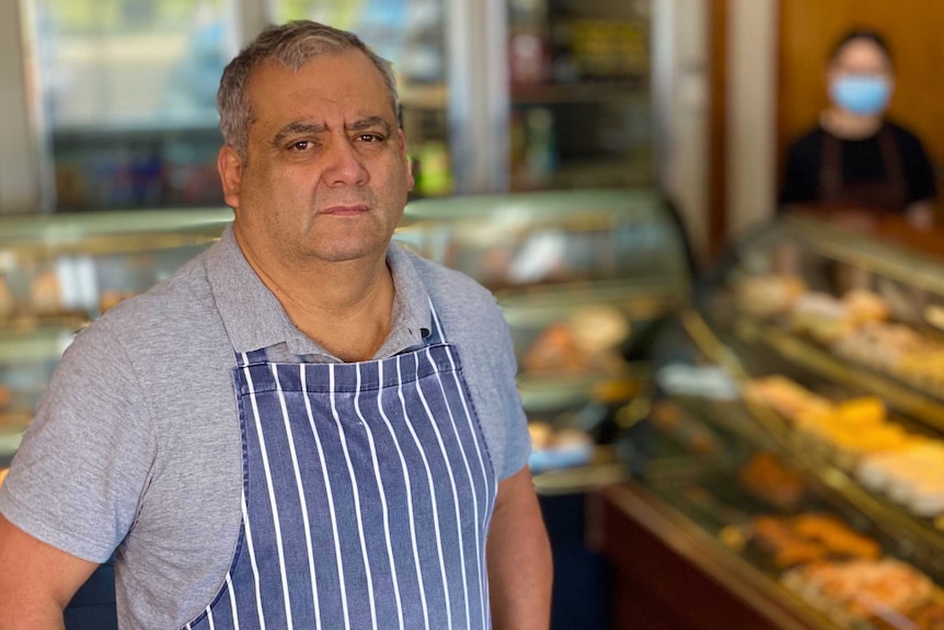 A man wearing an apron stands in front of a display of cakes in a cake shop.