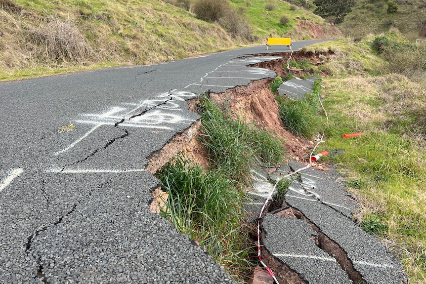A bitumen road which has subsided on the right hand side. Pieces of bitumen lie a metre below the road level with grass growing 