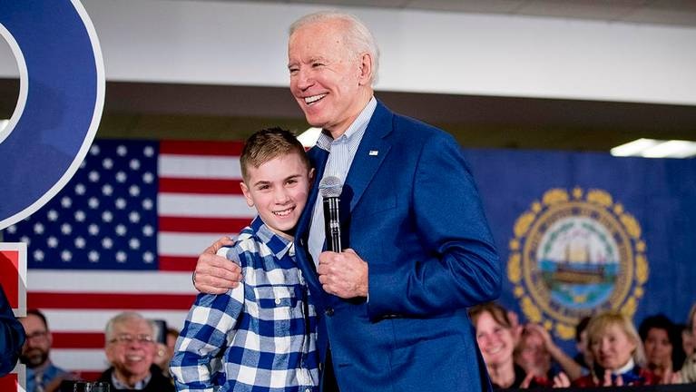 US President Elect Joe Biden hugs Brayden Harrington on stage in front of American flag.