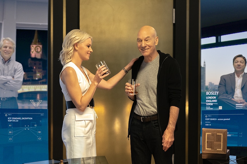 Elizabeth Banks stands with hand on shoulder of Sir Patrick Stewart, both hold glasses in a room with wall-sized video screens.