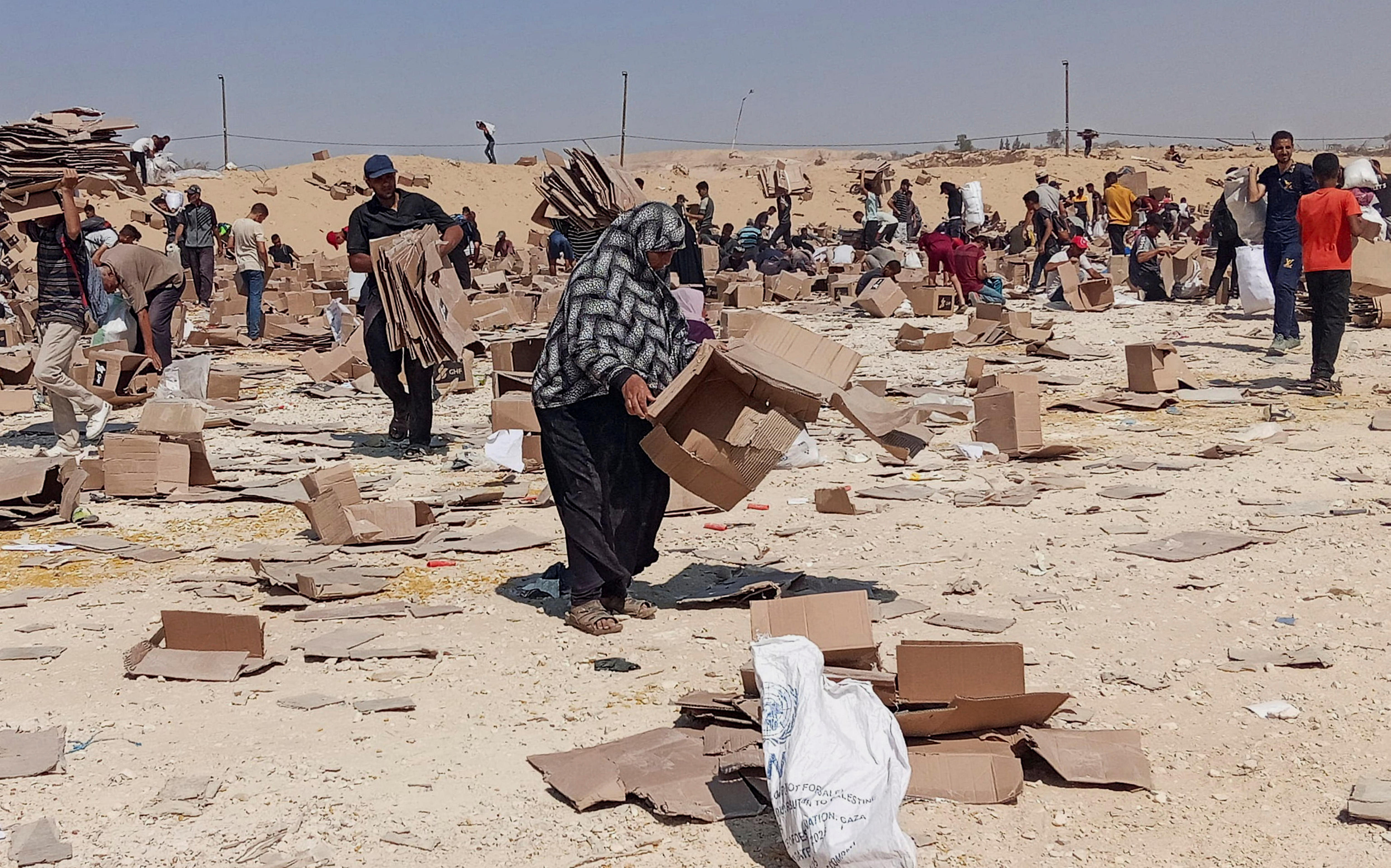 A woman picks up an empty cardboard box from the ground as a crowd looks through rubbish on dusty ground.