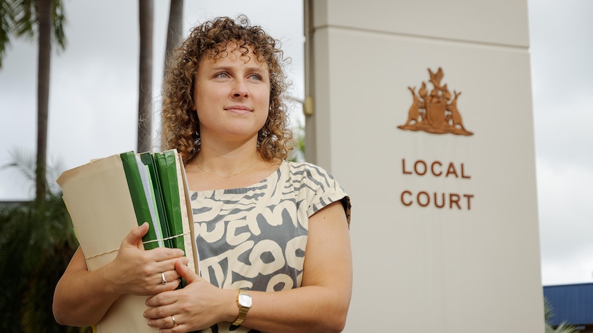 A woman with curly hair and a printed top holds a stack of paperwork outside Darwin local court.