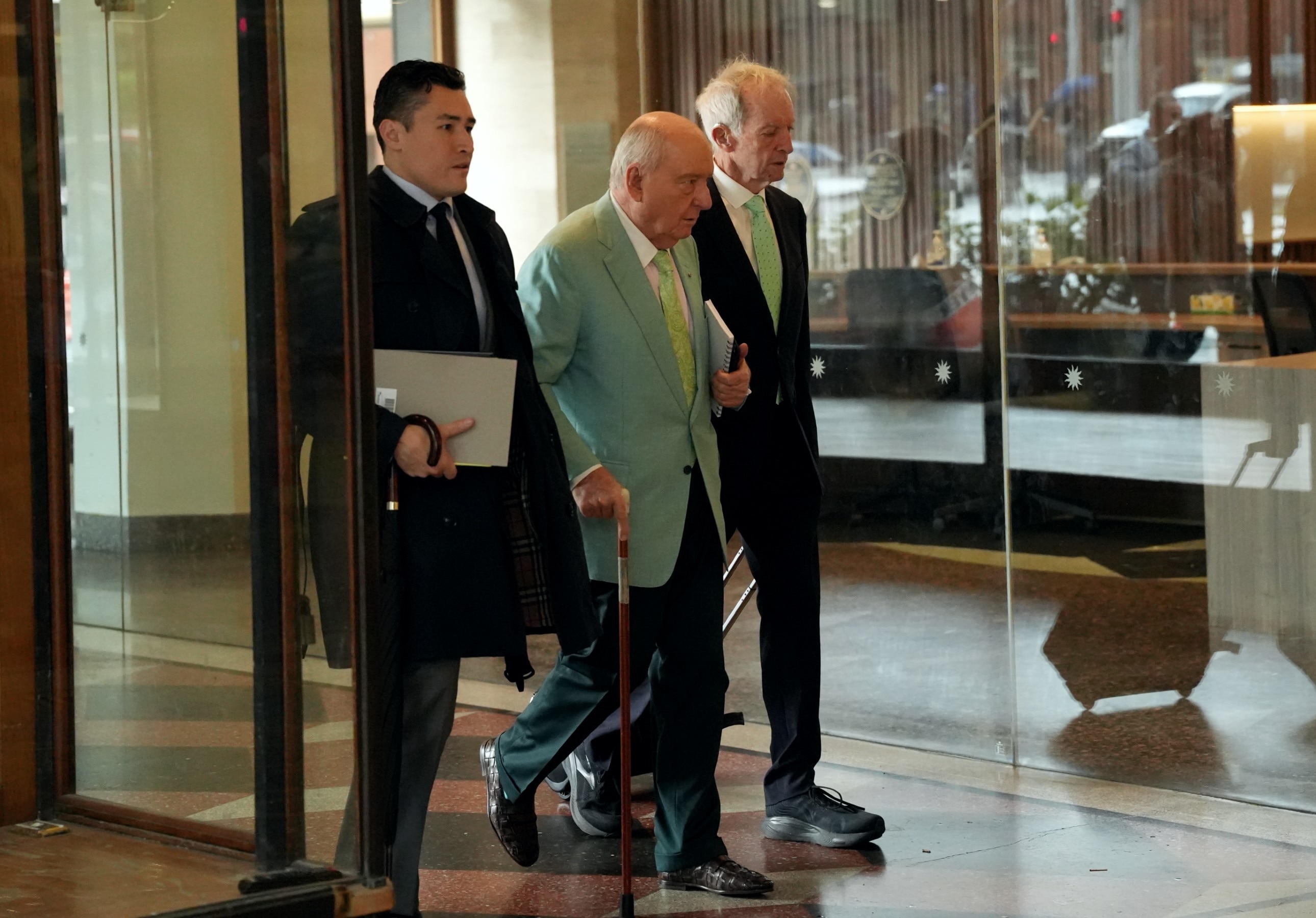 Man in teal suit jacket in street walking in foyer with men in suits on either side